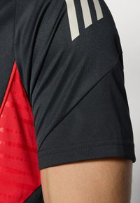 Svart sporttröja med röda detaljer, med diagonalt mönster, slät textur och tre vertikala vita ränder på axeln.