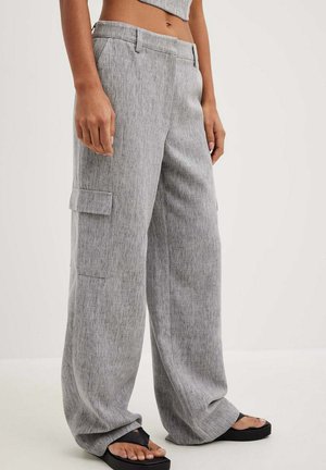 Kvinde iført løstsiddende grå cargobukser med sidelommer og sorte plateau-sandaler, stående mod en ensartet lys baggrund.