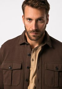 Bruine casual jas met twee borstzakken en zwarte knopen, gelaagd over een beige poloshirt. De stof lijkt textuur en zwaar.