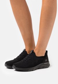 Černé sportovní boty s texturovaným pleteným svrškem, elastickým šněrováním, nízkým střihem kolem kotníku a polstrovanou černou podrážkou. Žádné výrazné loga nejsou viditelná.