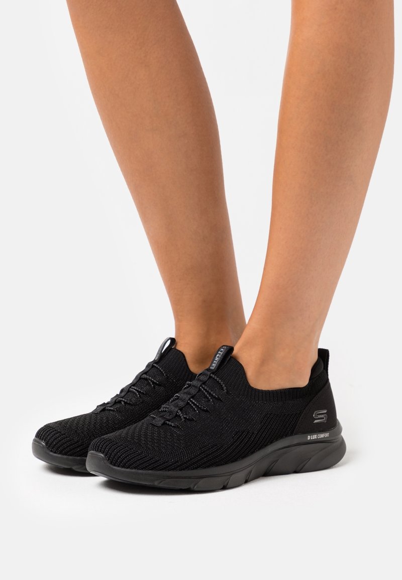 Černé sportovní boty s texturovaným pleteným svrškem, elastickým šněrováním, nízkým střihem kolem kotníku a polstrovanou černou podrážkou. Žádné výrazné loga nejsou viditelná.