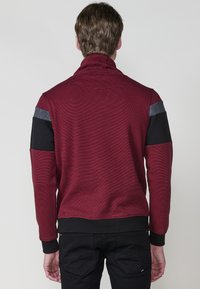 Burgundský pulover s vysokým golierom, ktorý má čierne a šedé akcentové panely na pleciach a textúrovanú látku.