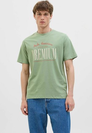 Zeleni pamučni T-shirt s printom "PREMIUM" u krem i burgundskom tonu. Kratki rukavi i okrugli izrez. Nositi uz plave traperice.