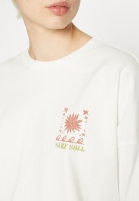 Rövid ujjú, fehér pamut póló, színes grafikai nyomattal, amelyen egy nap és a "SURF SOUL" felirat zöld, rózsaszín és narancssárga színekben látható.