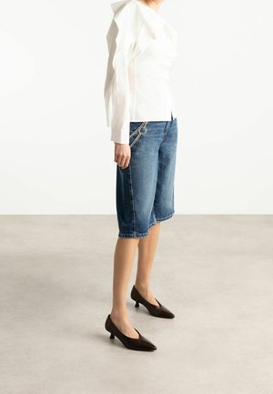 Woman wearing a white ruffled blouse, knee-length blue denim shorts with chain detail, and dark brown low-heeled pointed shoes.