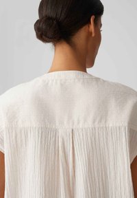 Blusa leggera color crema con una finitura testurizzata, caratterizzata da maniche corte, un dettaglio plissè posteriormente e un collo rotondo.