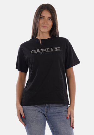 Donna con capelli castani lisci che indossa una maglietta nera a maniche corte con la scritta "GAELLE" in strass e jeans blu, in piedi davanti a uno sfondo bianco.