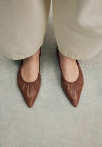 Barna bőralacsony sarkú cipők hegyes orral és ráncolt elasztikus részletekkel a nyílás körül, laza krémszínű nadrággal viselve.
