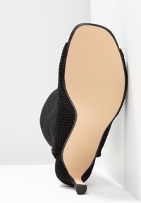 Bottine noire texturée à talon haut avec bout pointu, semelle beige et talon fin, posée contre un angle blanc.