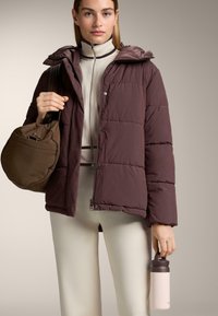 Bordeaux polstret jakke med hætte, lynlåslukning og trykknapper. Beige tætsiddende top, lyse bukser, en brun taske og en lyserød vandflaske.