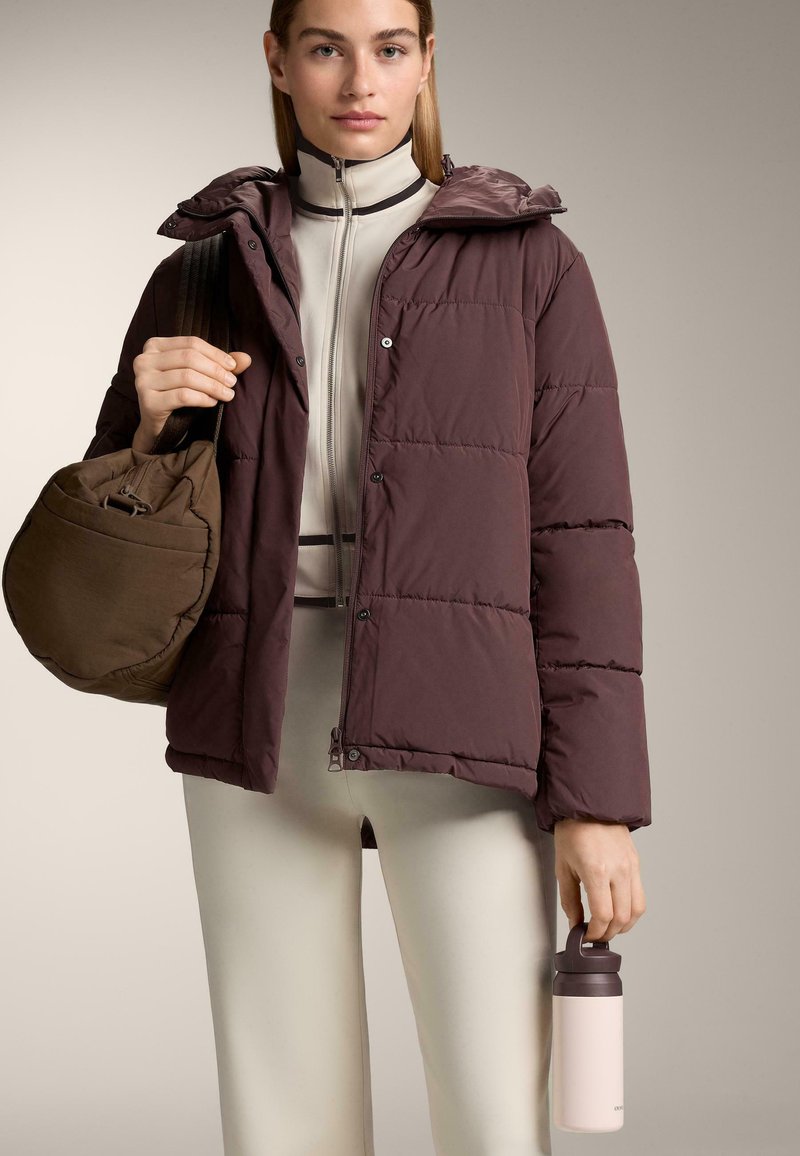 Bordeaux polstret jakke med hætte, lynlåslukning og trykknapper. Beige tætsiddende top, lyse bukser, en brun taske og en lyserød vandflaske.