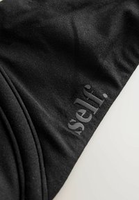 Zwarte stof die een gladde textuur toont, met daarin de verhoogde letterbranding "self." in een contrasterende donkere afwerking. Gebogen randen zijn zichtbaar.