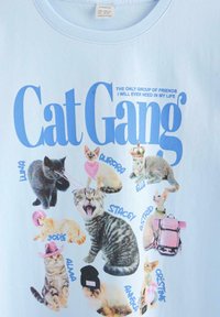 Lyseblå T-shirt-tryk med ni katte, navngivet og iført tilbehør, med titlen "Kattegeng" og en sætning om venskab ovenover.