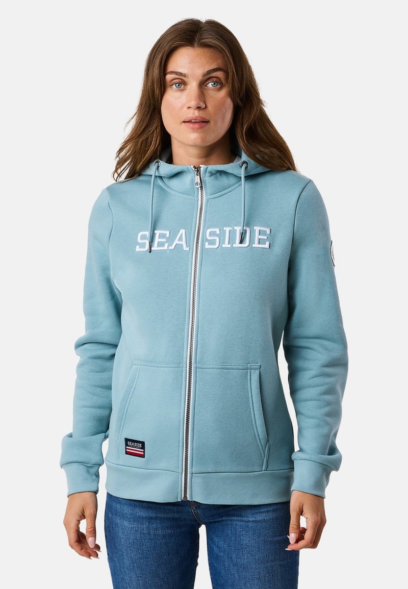 Lichtblauwe rits hoodie van zacht materiaal. Voorzien van "SEA SIDE" in witte letters, voorzakken en verstelbare trekkoorden op de capuchon.