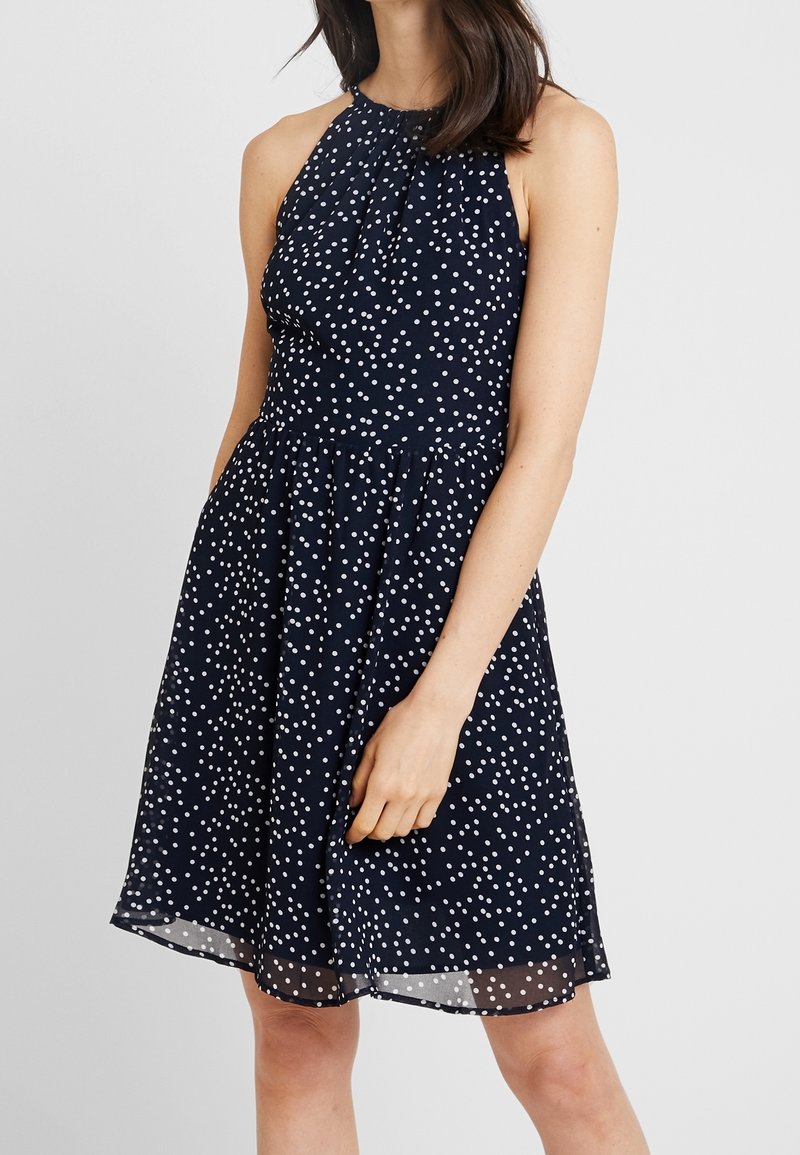 Femme portant une robe bleu marine sans manches à col licou avec des pois blancs, debout, tenant délicatement la jupe d'une main.