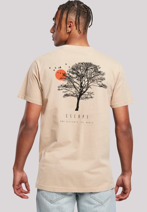 Man in een beige T-shirt met een zwart boom, een oranje zon en vogels grafiek en de tekst "ESCAPE en ontdek de wereld" op de achterkant.