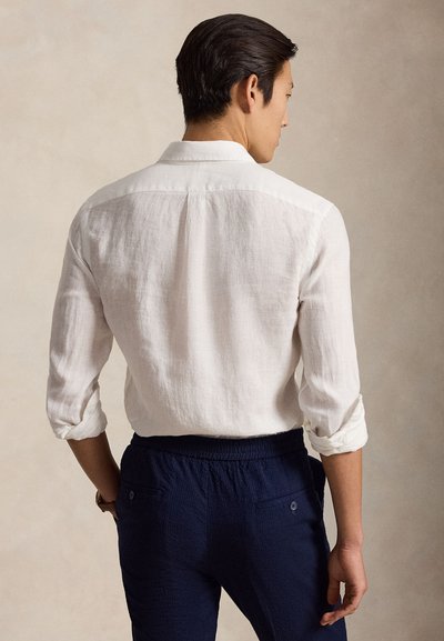 Άνδρας που φοράει λευκό πουκάμισο με μακριά μανίκια, γυρισμένα στο ύψος των αγκώνων, και σκούρο μπλε παντελόνι, στέκεται με τα χέρια στις τσέπες, γυρισμένος προς τα πίσω.