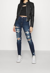 Svart lädermotorcykeljacka, kort svart topp, slitna blå skinnyjeans och vita sneakers. Jeansen har revor och lappar.