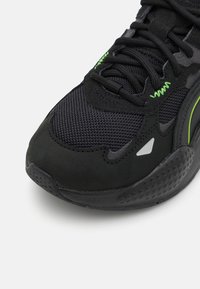 Musta urheilukenkä, jossa on verkkopinta vihreillä yksityiskohdilla, mokkamateriaalilla päällystyksiä ja teksturoitu kumipohja geometrisilla kuvioilla.