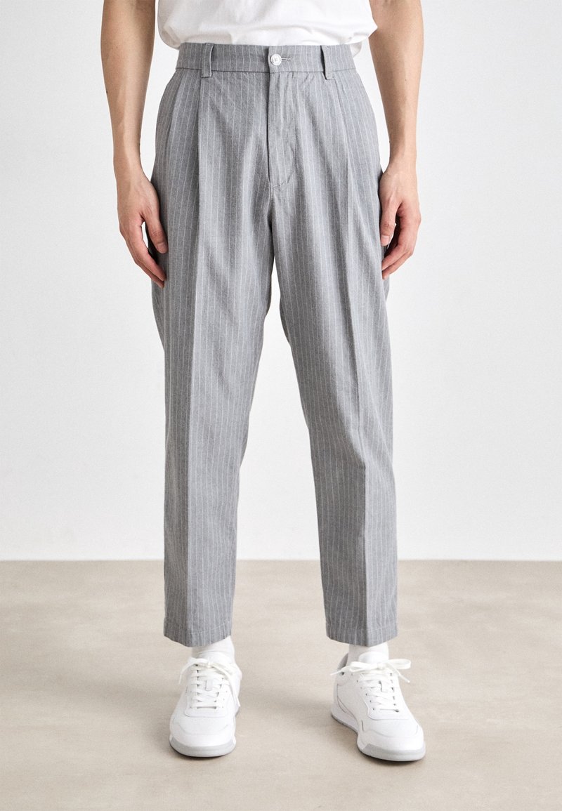 Homme portant un pantalon gris à fines rayures, des chaussettes blanches, des baskets blanches et une chemise blanche rentrée, debout sur un sol neutre.