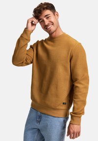 Sudadera amarilla mostaza con tejido texturizado, cuello redondo, puños acanalados y un patrón geométrico. Combinada con jeans de tono azul claro.