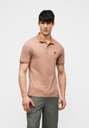 Férfi, világosbarna rövid ujjú galléros pólóban, amelyen apró sárga madár logó látható a mellkason, szürke nadrággal párosítva.