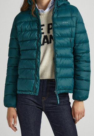 Teal pufferjack met een horizontaal gestikt patroon, ritssluiting, een vaste capuchon en elastische manchetten, gedragen over een crème trui en donkere spijkerbroek.