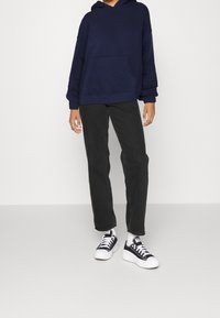 Felpa blu navy con tasca frontale, abbinata a pantaloni neri a gamba dritta e sneakers bianco-neri, con suole e lacci bianchi.
