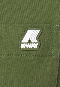 Tela de color verde oliva con textura acanalada, que presenta un parche con el logo de K-WAY en blanco. Diseño sencillo, detalles mínimos y costuras visibles.