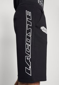 Shorts noirs avec le grand texte blanc "LACOSTE" verticalement sur le côté et un plus petit logo Lacoste près de la poche sur la jambe d'une personne.