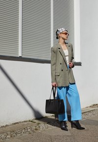Blazer doppiopetto verde chiaro su una maglietta bianca, abbinato a pantaloni ampi blu brillante e stivali neri con tacco alto, mentre tiene una borsa a tracolla nera.