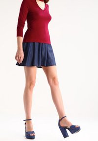 Punainen pitkähihainen paita V-pääntiellä, yhdistettynä tummansinisestä denimsateesta valmistettuun flare-hameeseen ja tummansinisiin korotettuihin sandaaleihin nilkkaremmillä.