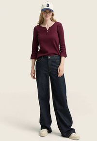 Maron rød langærmet henley-shirt, vidtbenede mørke denimjeans, hvide sneakers og en hvid baseballkasket med en blå accent på skyggen.