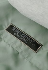 Lys grøn sportstøjstof med en glat tekstur. Har et sort mærke med "SUPERDRY NYC" og "SPORTSWEAR" i hvid syning.