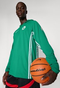 Pikk mees kannab rohelise Boston Celticsi kapuutsi ja hoiab käes oranži korvpalli, seistes heleda ühtlase tausta ees.