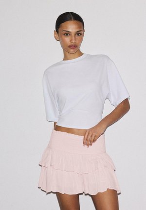 Kobieta o ciemnych włosach związanych w gładki kok, ubrana w biały krótki top z krótkim rękawem oraz jasnoróżową, warstwową mini spódnicę na jednolitym tle.