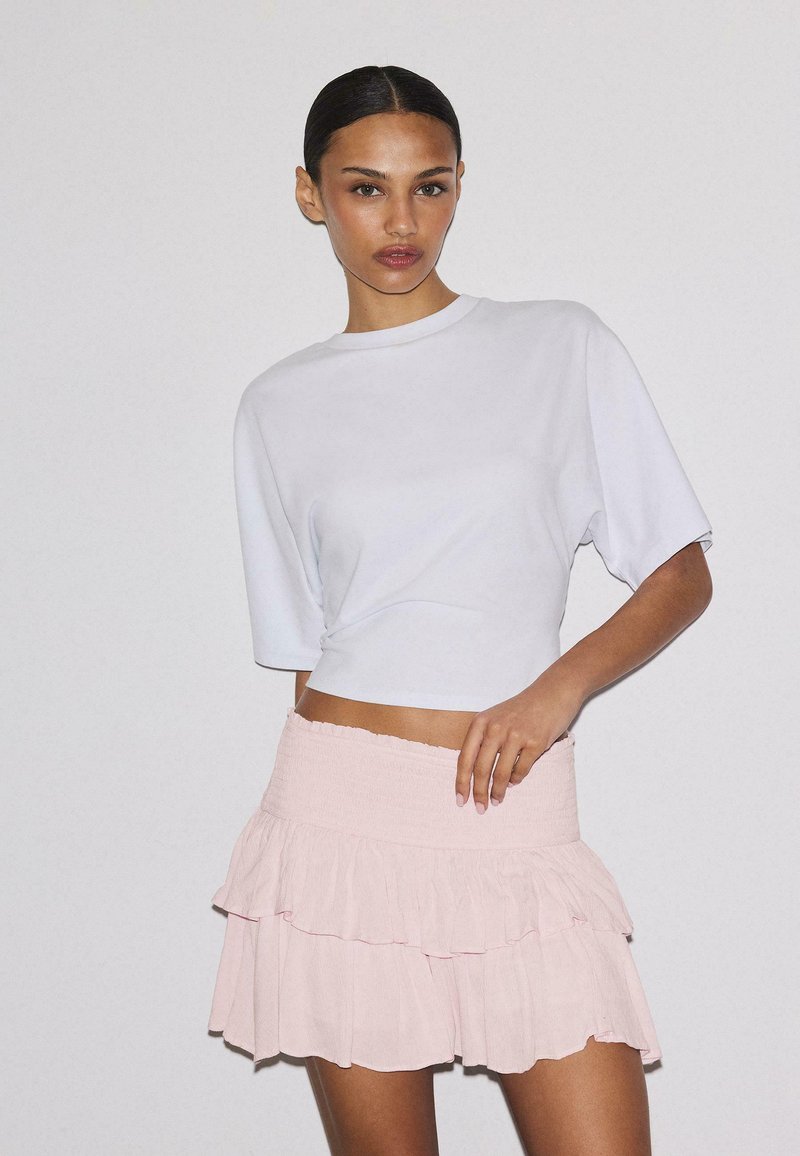 Femme aux cheveux foncés coiffés en chignon élégant, portant un haut blanc à manches courtes et court, ainsi qu'une mini-jupe à volants rose pâle, sur un fond uni.