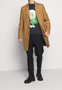 Płaszcz z owczej wełny w kolorze wielbłądzim z klapami z wycięciem, trzema guzikami i jedną kieszenią na piersi, noszony na czarnej bluzie z nadrukiem i ciemnych spodniach.