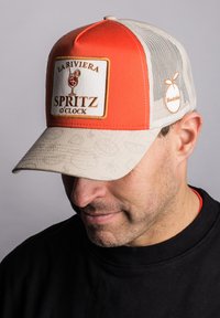 Trucker-Cap mit einer leuchtend orangefarbenen Vorderseite und beigem Netzrücken. Verfügt über ein quadratisches Aufnäher mit der Aufschrift "LA RIVIERA SPRITZ O'CLOCK" und einen strukturierten Schirm.