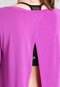 Lila, lätt top med öppning i ryggen och korta ärmar, med en jämn textur och avslappnad passform som blottar en svart linne under.