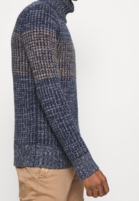 Pull en tricot bleu marine avec des accents beiges, dotée d'un col montant, d'un motif texturé et de poignets côtelés. Le matériau semble épais et chaud.