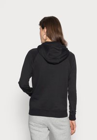 Felpa nera con cappuccio, maniche lunghe, polsini e orlo a coste. Materiale morbido, vestibilità comoda, cappuccio visibile sul retro. Pantaloni grigi visibili.