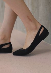 Sorte suedes ballettflater med spiss tå og åpne sidesskjæringer, hviler på en glatt, lys rosa overflate.