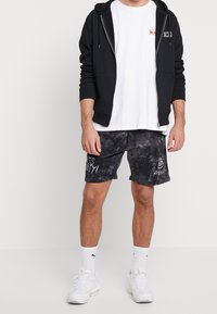 Felpa nera con zip, t-shirt bianca, pantaloni corti neri tie-dye con testo grafico e scarpe da ginnastica bianche. Il tessuto sembra morbido; design casual.