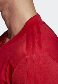 Chemise de sport rouge avec un col en V, dotée d'un tissu texturé et de trois bandes verticales sur l'épaule. Esthétique générale lisse.