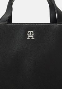 Sac en cuir noir avec une texture légèrement granuleuse et un logo en métal argenté "TH" sur le devant, équipé de deux poignées supérieures pour le transport.