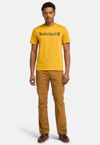 Keltainen puuvillapaita, jossa on "Timberland"-logo tummanharmaana, yhdistettynä ruskeisiin housuihin ja viininpunaisiin kenkiin. Yksinkertainen muotoilu, rento istuvuus.
