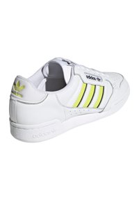 Fehér bőr sneaker, három fekete és neon sárga csíkkal, kerek orral, párnázott nyakrésszel és texturált talppal, amelyen a saroknál a márkajelzés található.