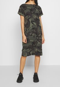 Robe en coton à imprimé camouflage dans les tons de vert foncé et noir, à manches courtes, coupe décontractée, longueur genoux, associée à des baskets noires.