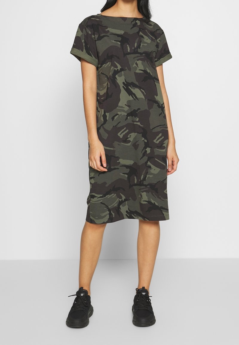 Robe en coton à imprimé camouflage dans les tons de vert foncé et noir, à manches courtes, coupe décontractée, longueur genoux, associée à des baskets noires.